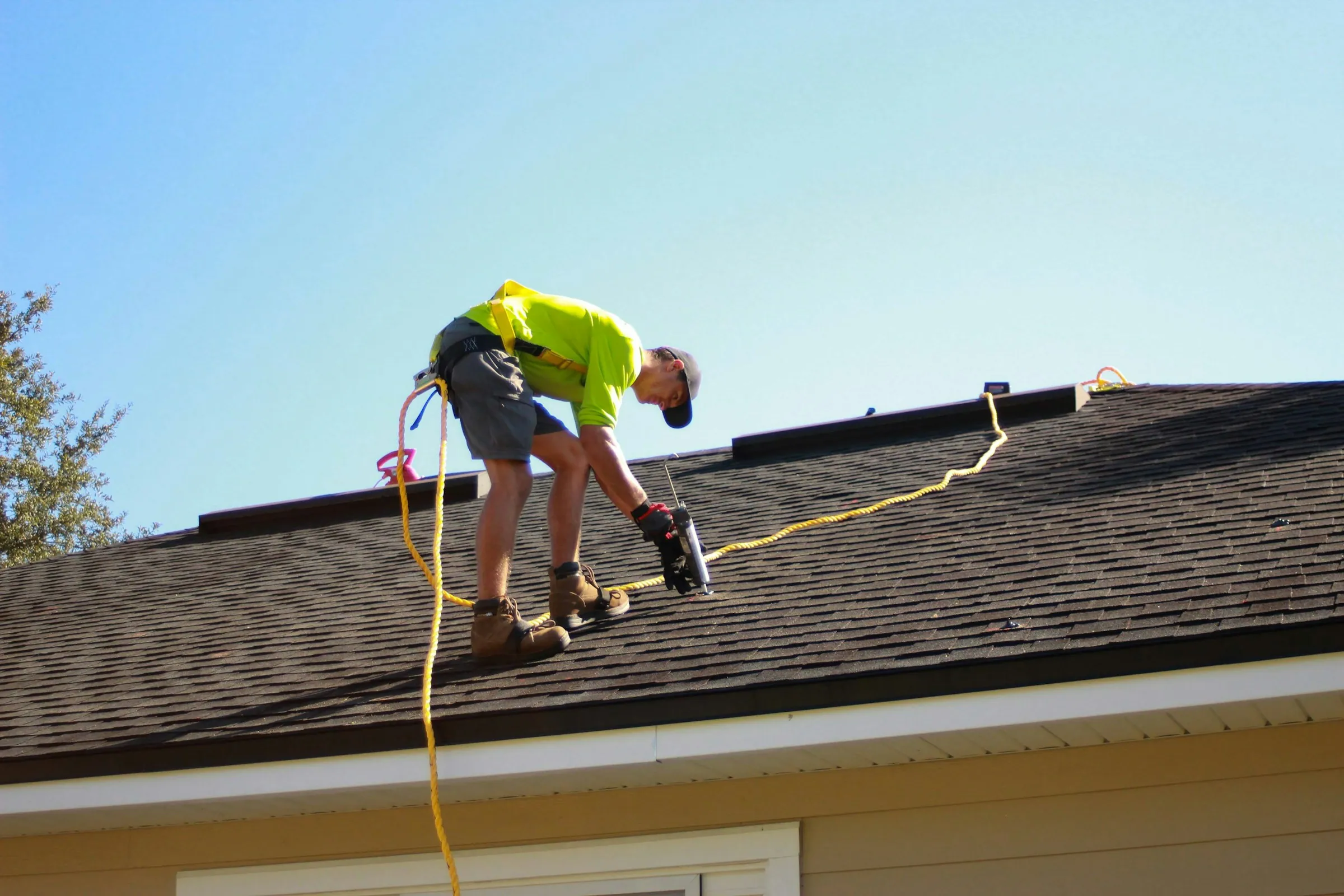 A roofer working on a shingled roof under a clear sky